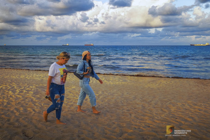 Two women walking on Fort Lauderdale Beach sand with cargo container ships in the background offshore