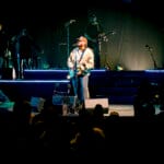 James Arthur, wearing a hat and jacket, sings passionately while playing an acoustic-electric guitar on a dramatically lit blue stage at Yaamava' Theater, with his band in the background.