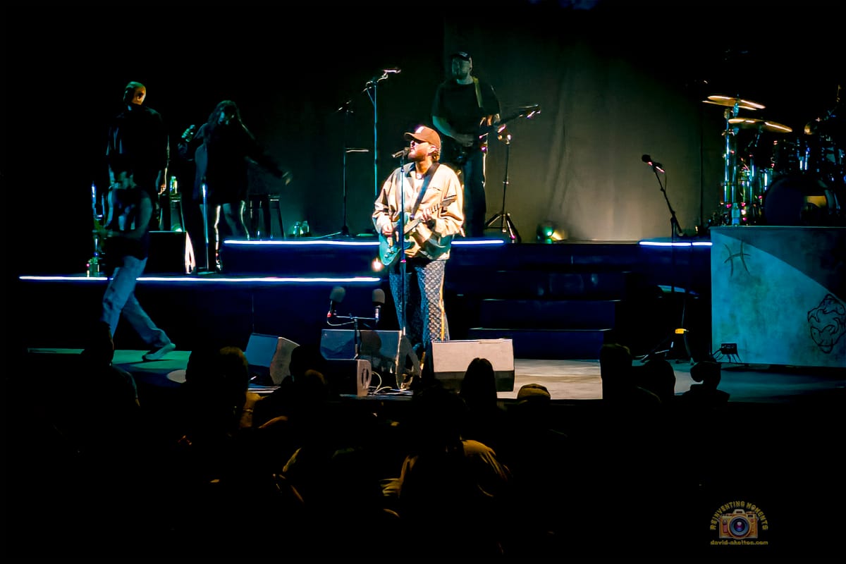 James Arthur, wearing a hat and jacket, sings passionately while playing an acoustic-electric guitar on a dramatically lit blue stage at Yaamava' Theater, with his band in the background.