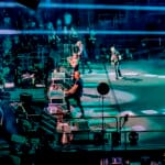 Wide shot of the rock band Creed performing on a large, blue-lit stage in a packed arena, with singer Scott Stapp on the left and a massive speaker array hanging from the ceiling.