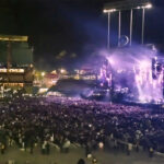 A wide night shot of Billy Joel performing on a brightly lit stage at a sold-out Dodger Stadium, with thousands of fans in the crowd holding up their phone lights.