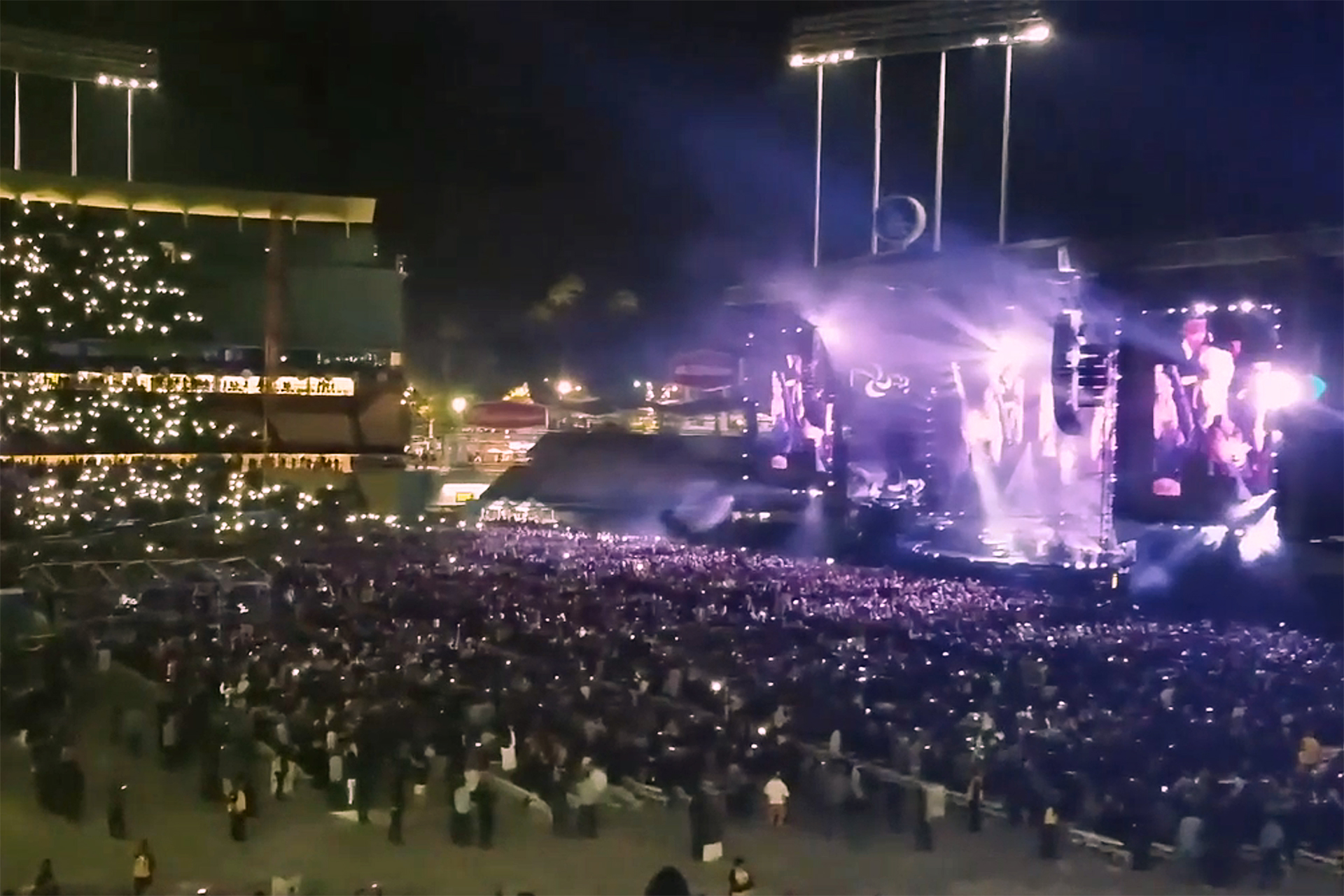 A wide night shot of Billy Joel performing on a brightly lit stage at a sold-out Dodger Stadium, with thousands of fans in the crowd holding up their phone lights.