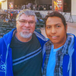 A smiling selfie of two men in blue hoodies at the Sleep Train Amphitheatre, with the concert stage and seating visible in the background before a show.