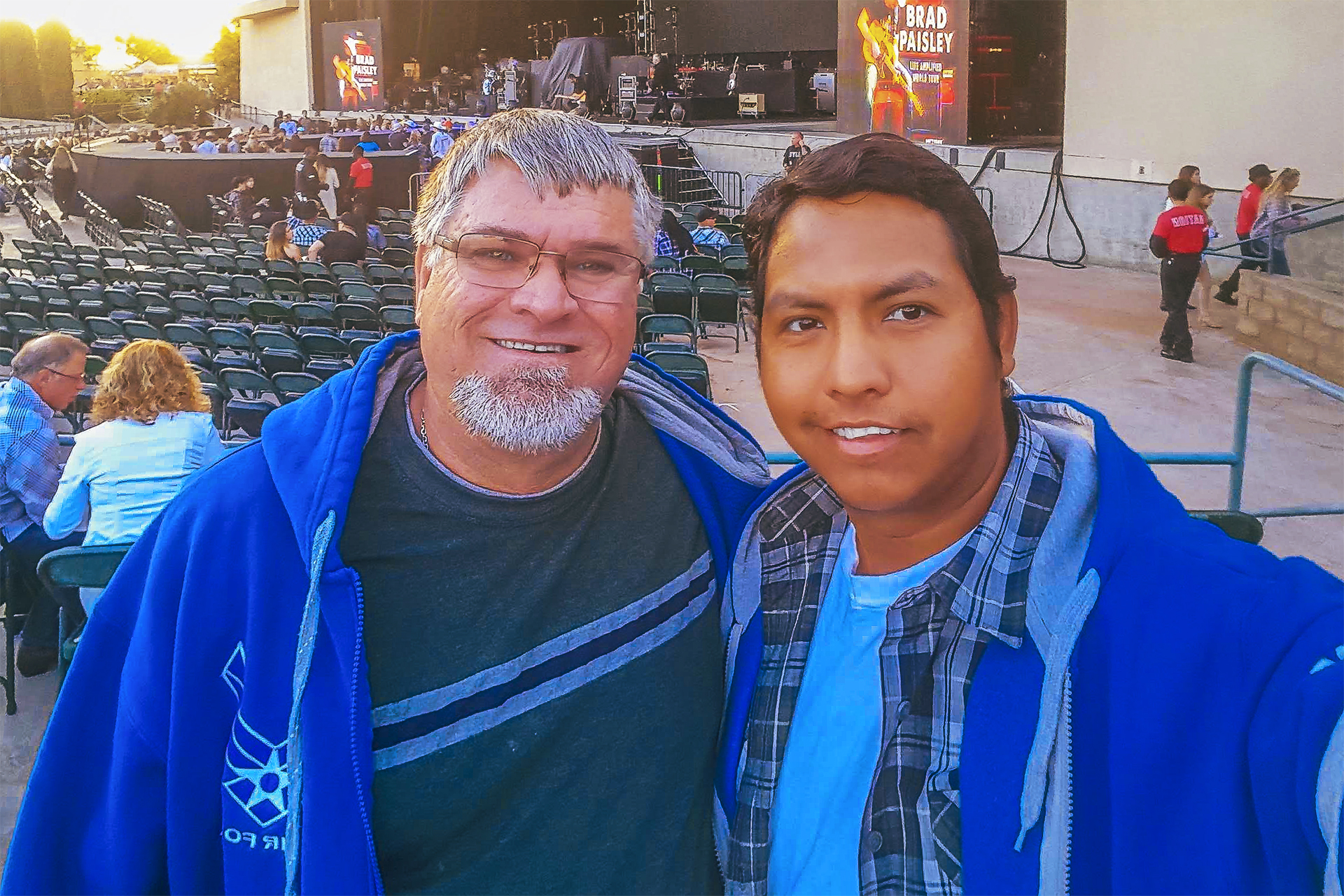 A smiling selfie of two men in blue hoodies at the Sleep Train Amphitheatre, with the concert stage and seating visible in the background before a show.