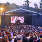 A wide shot from the audience of the Bryan Adams "Get Up" tour at the Greek Theatre in Los Angeles at dusk. The stage's main screen shows a black and white image of the artist, while a large crowd fills the outdoor amphitheater's seats.