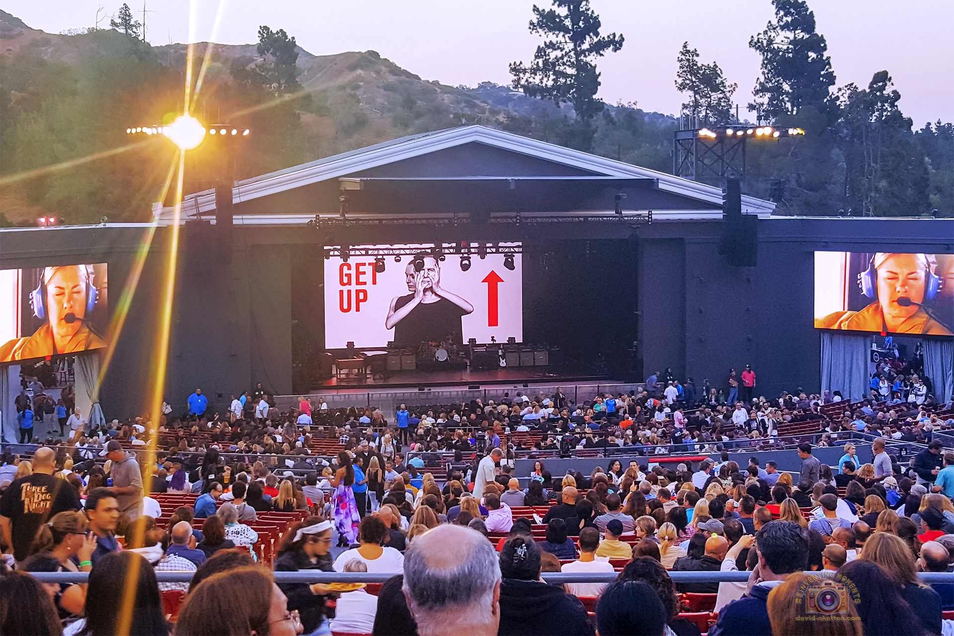 A wide shot from the audience of the Bryan Adams "Get Up" tour at the Greek Theatre in Los Angeles at dusk. The stage's main screen shows a black and white image of the artist, while a large crowd fills the outdoor amphitheater's seats.