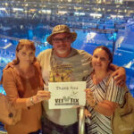 A man and two women smiling for a photo from the upper deck of STAPLES Center, holding a "Thank You Vet Tix" sign, with the 360-degree concert stage visible below.