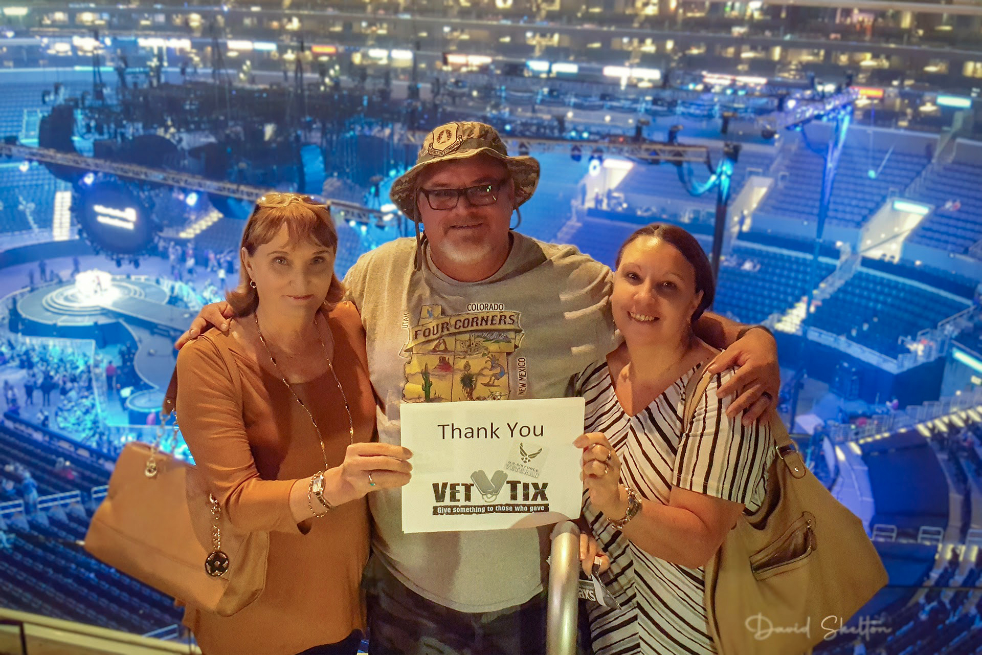 A man and two women smiling for a photo from the upper deck of STAPLES Center, holding a "Thank You Vet Tix" sign, with the 360-degree concert stage visible below.