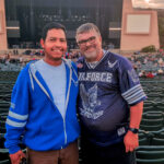 Two men smiling for a photo in front of their seats at the Sleep Train Amphitheatre, with the large concert stage and empty venue seats in the background before a show.