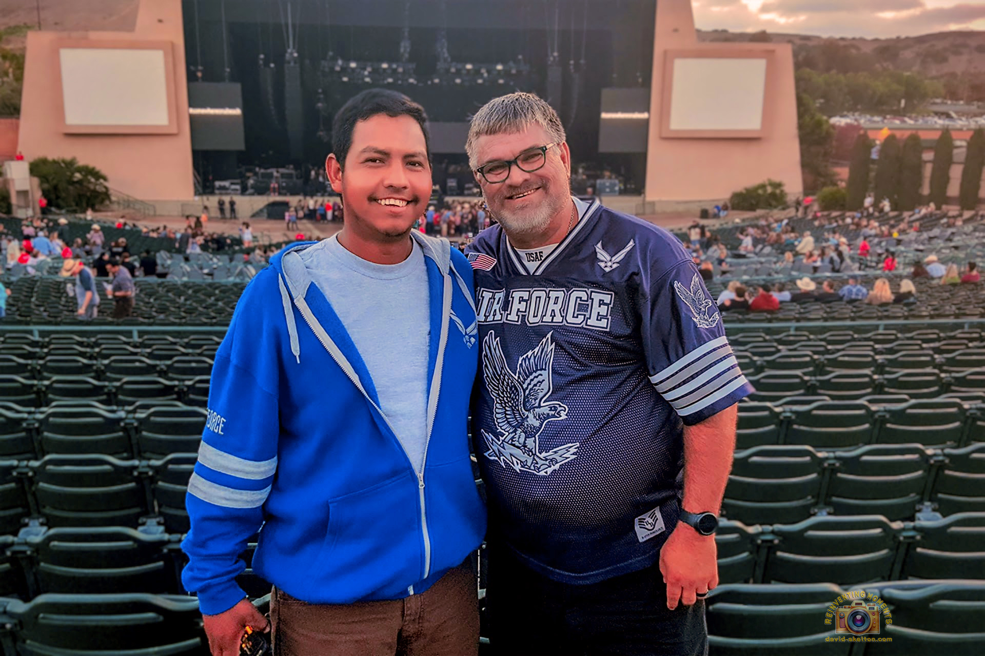 Two men smiling for a photo in front of their seats at the Sleep Train Amphitheatre, with the large concert stage and empty venue seats in the background before a show.
