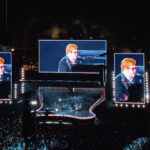 A wide shot of Elton John's "Farewell Yellow Brick Road" concert at Dodger Stadium at night. Three large screens dominate the view, showing close-ups of Elton John in his iconic glasses playing the piano for a massive crowd.