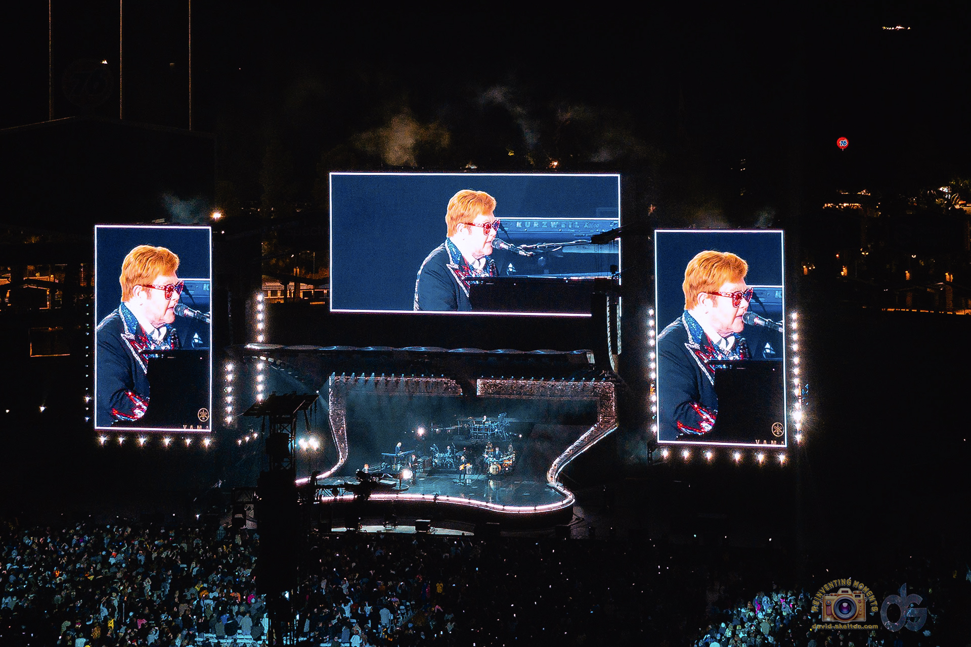 A wide shot of Elton John's "Farewell Yellow Brick Road" concert at Dodger Stadium at night. Three large screens dominate the view, showing close-ups of Elton John in his iconic glasses playing the piano for a massive crowd.
