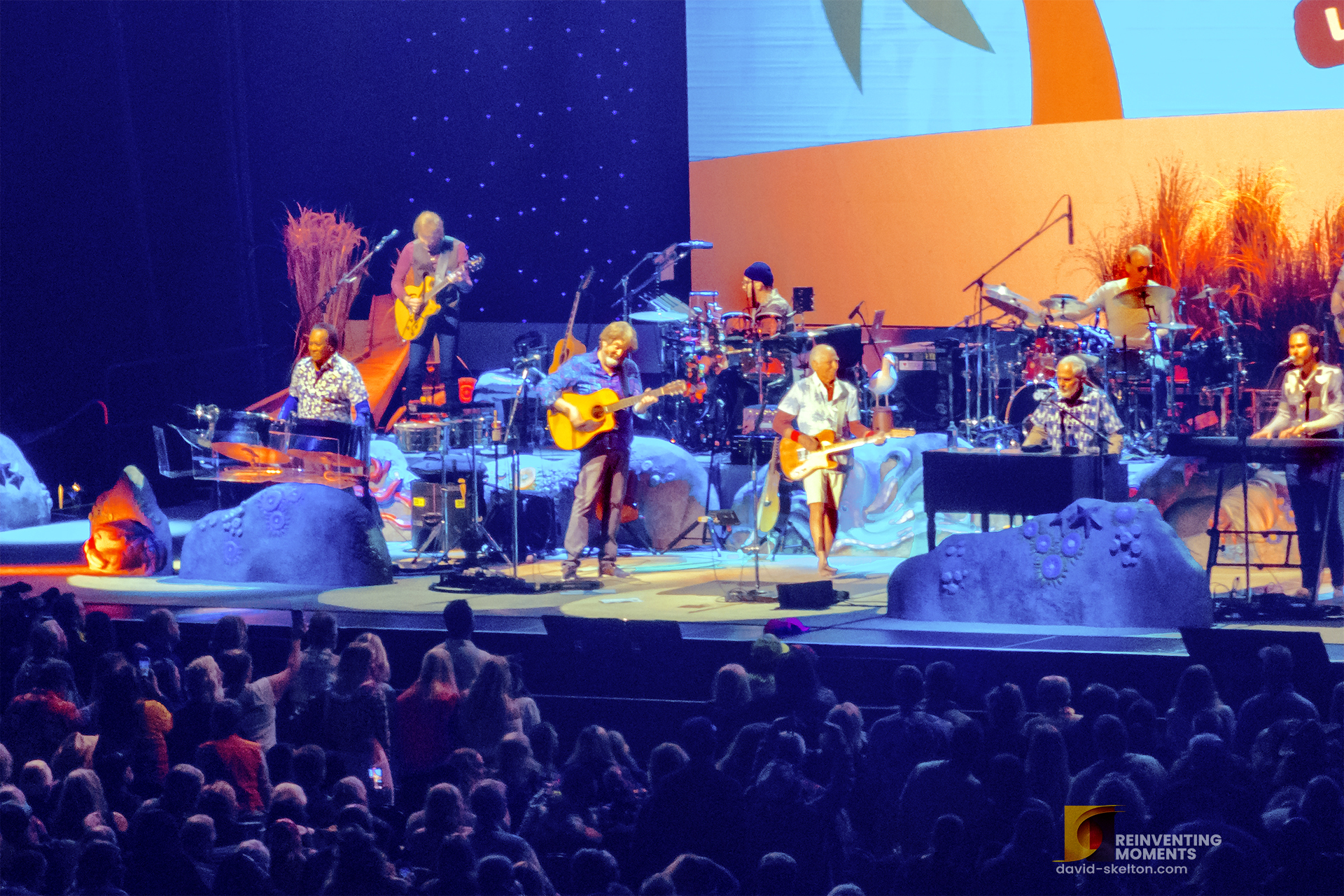 Jimmy Buffett and the Coral Reefer Band performing live on a tropically decorated stage with blue and orange lighting in front of a large concert audience at Acrisure Arena.