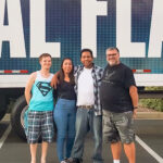 A group of four friends smiling for a photo in a parking lot in front of a large tour truck before a concert.