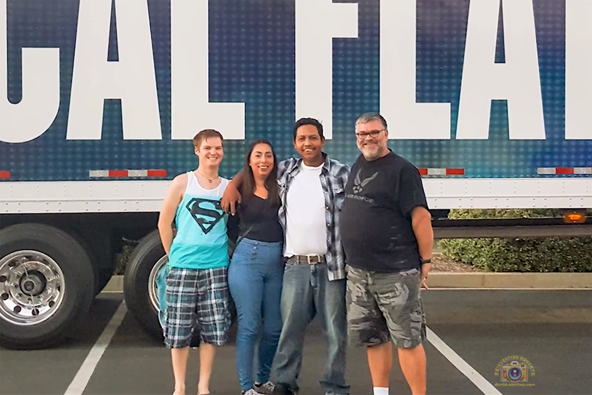 A group of four friends smiling for a photo in a parking lot in front of a large tour truck before a concert.
