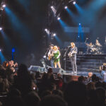 A wide shot of Reba McEntire, Brooks & Dunn performing together on stage during their Las Vegas residency. Reba, in a sparkling green outfit, stands between Kix Brooks and Ronnie Dunn as they play guitar for a large audience.