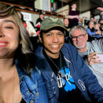 A smiling group selfie of a woman and two men in the audience seats at the Honda Center for a Bryan Adams concert. The man in the middle wears a hat, and the man on the right is waving at the camera.