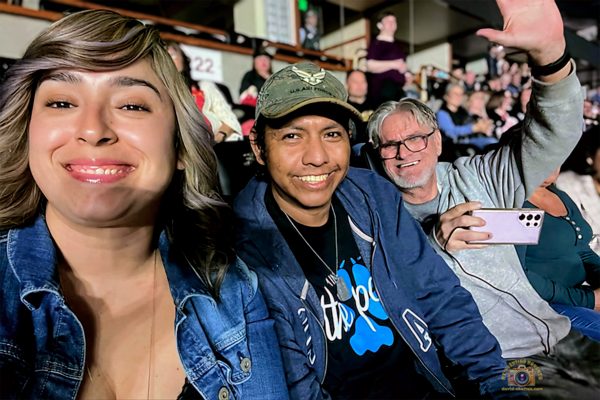 A smiling group selfie of a woman and two men in the audience seats at the Honda Center for a Bryan Adams concert. The man in the middle wears a hat, and the man on the right is waving at the camera.