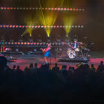 Carly Pearce singing and playing guitar on stage at the Pacific Amphitheatre during her 2025 Hummingbird Tour at the OC Fair. The stage is lit with colorful lights, and the audience is visible in silhouette.