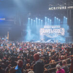 A wide-angle view from the stands of the packed Acrisure Arena during the Dierks Bentley concert, showing the stage illuminated in blue light with the "Gravel & Gold Tour" logo on the screen.