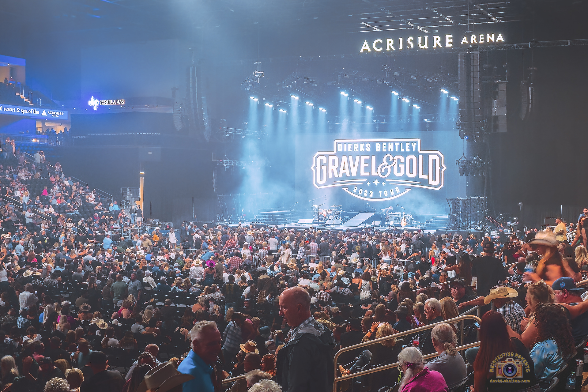 A wide-angle view from the stands of the packed Acrisure Arena during the Dierks Bentley concert, showing the stage illuminated in blue light with the "Gravel & Gold Tour" logo on the screen.