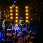 James Taylor and band performing live on stage with blue and orange lighting