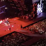 Kenny Chesney singing "Summertime" close-up on the extended stage at SoFi Stadium with large video screens behind him displaying live footage