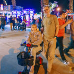 A man stands next to his mother, who is seated on a mobility scooter, smiling for a photo at the Riverside County National Date Festival at night. The background is filled with the bright, colorful lights of carnival rides and other fairgoers.