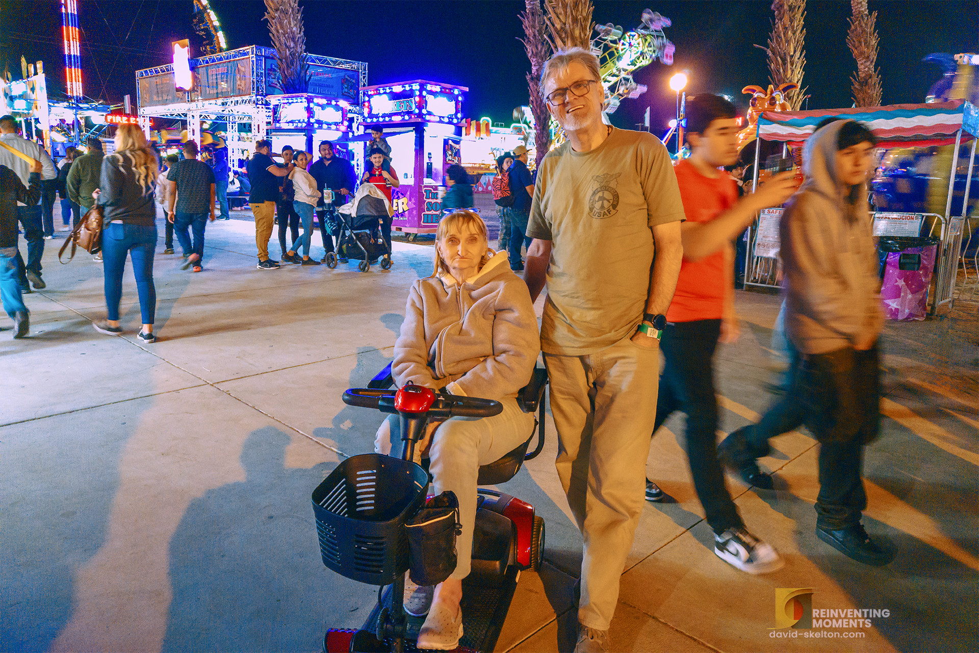 A man stands next to his mother, who is seated on a mobility scooter, smiling for a photo at the Riverside County National Date Festival at night. The background is filled with the bright, colorful lights of carnival rides and other fairgoers.