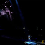 Tim McGraw singing "Highway Don't Care" with an outstretched hand, illuminated by a spotlight at the end of the stage at Kia Forum in Los Angeles during his Standing Room Only Tour. A video of McGraw plays behind him.