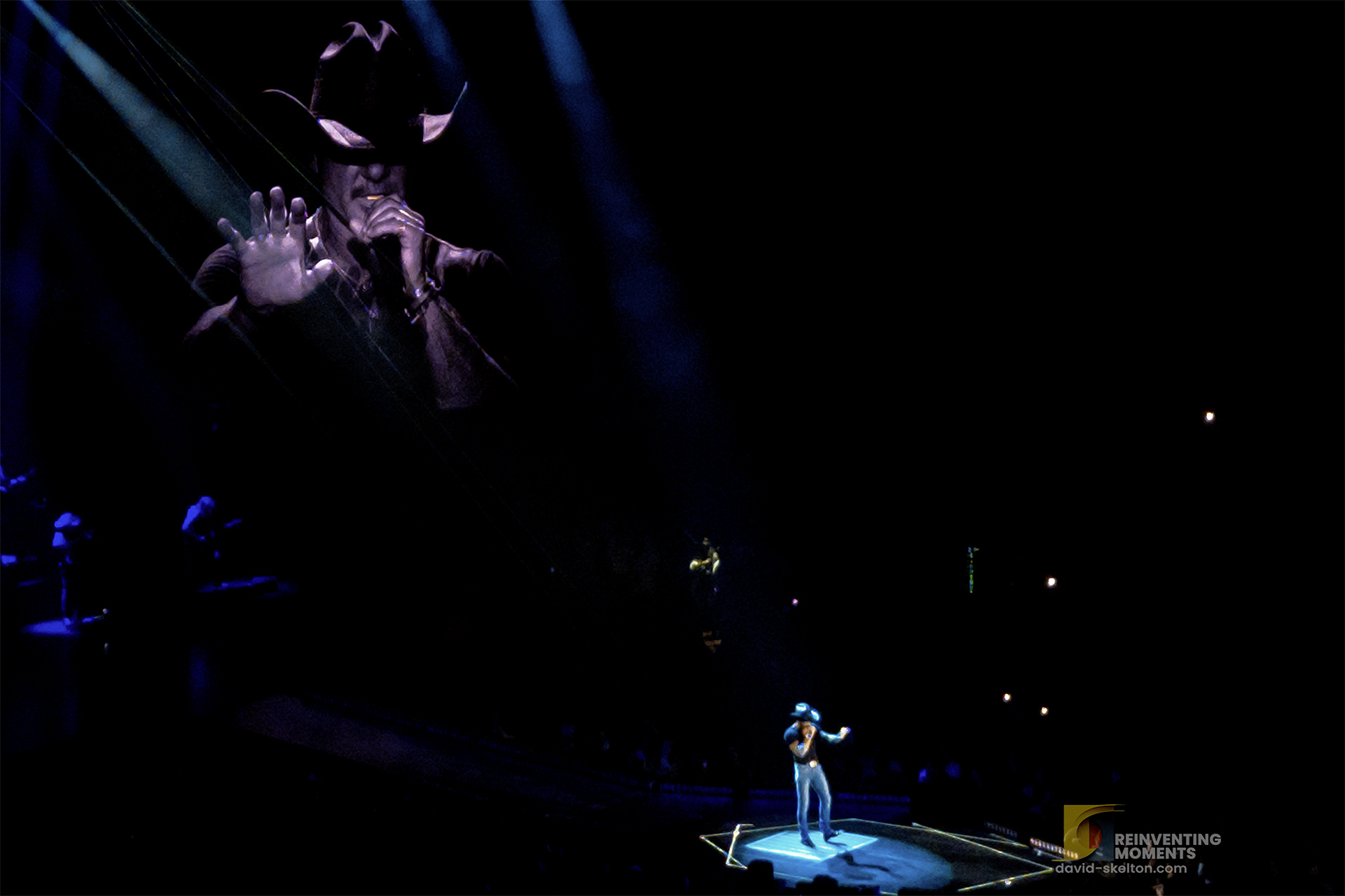 Tim McGraw singing "Highway Don't Care" with an outstretched hand, illuminated by a spotlight at the end of the stage at Kia Forum in Los Angeles during his Standing Room Only Tour. A video of McGraw plays behind him.
