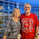 A man in a red Fire Rescue t-shirt stands with his arm around his mother on the brightly lit interior concourse of an arena at night.