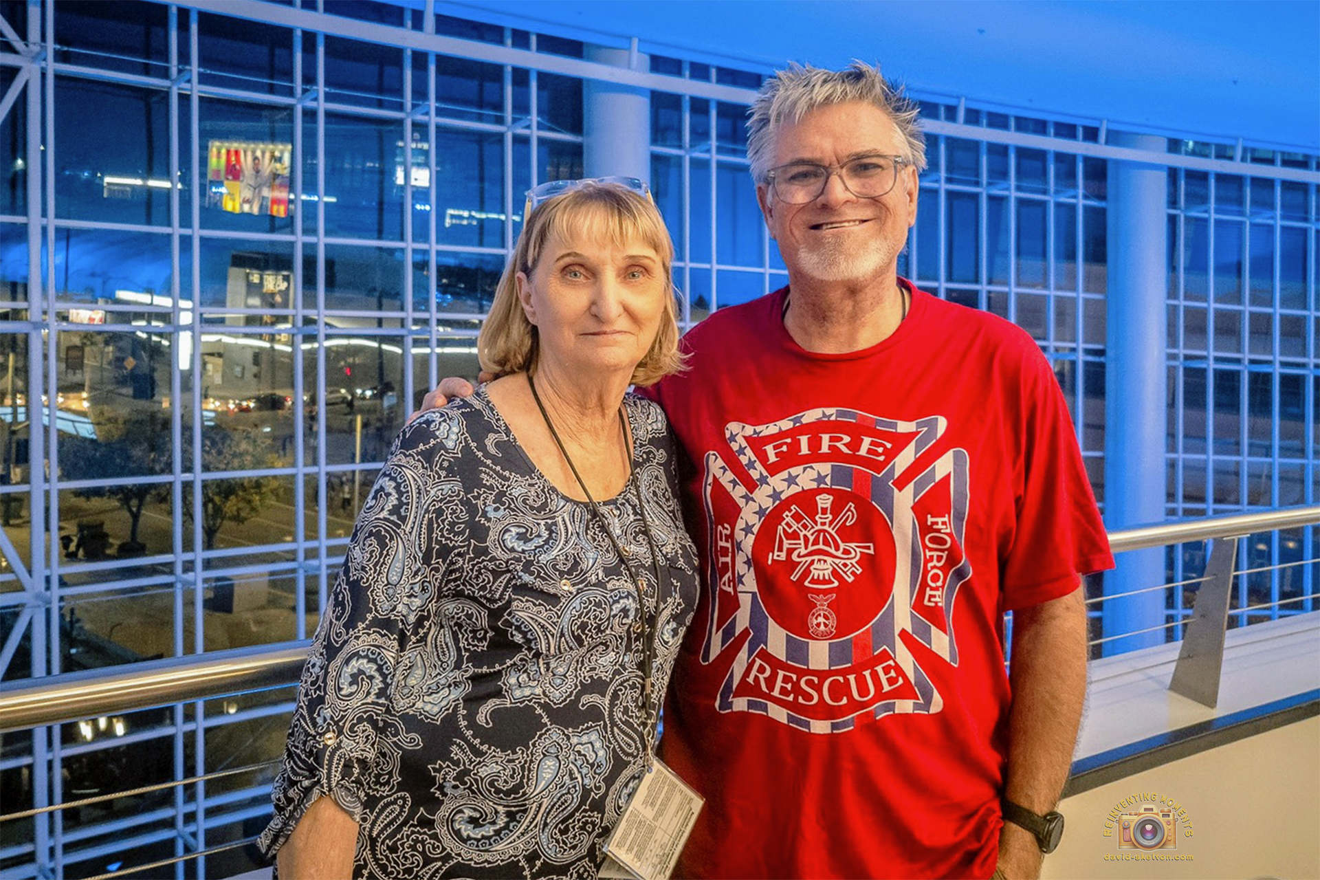 A man in a red Fire Rescue t-shirt stands with his arm around his mother on the brightly lit interior concourse of an arena at night.