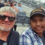 A man with white hair and sunglasses smiles next to his husband, who is wearing a plaid shirt and a baseball cap, sitting in the stands of a large, modern stadium.
