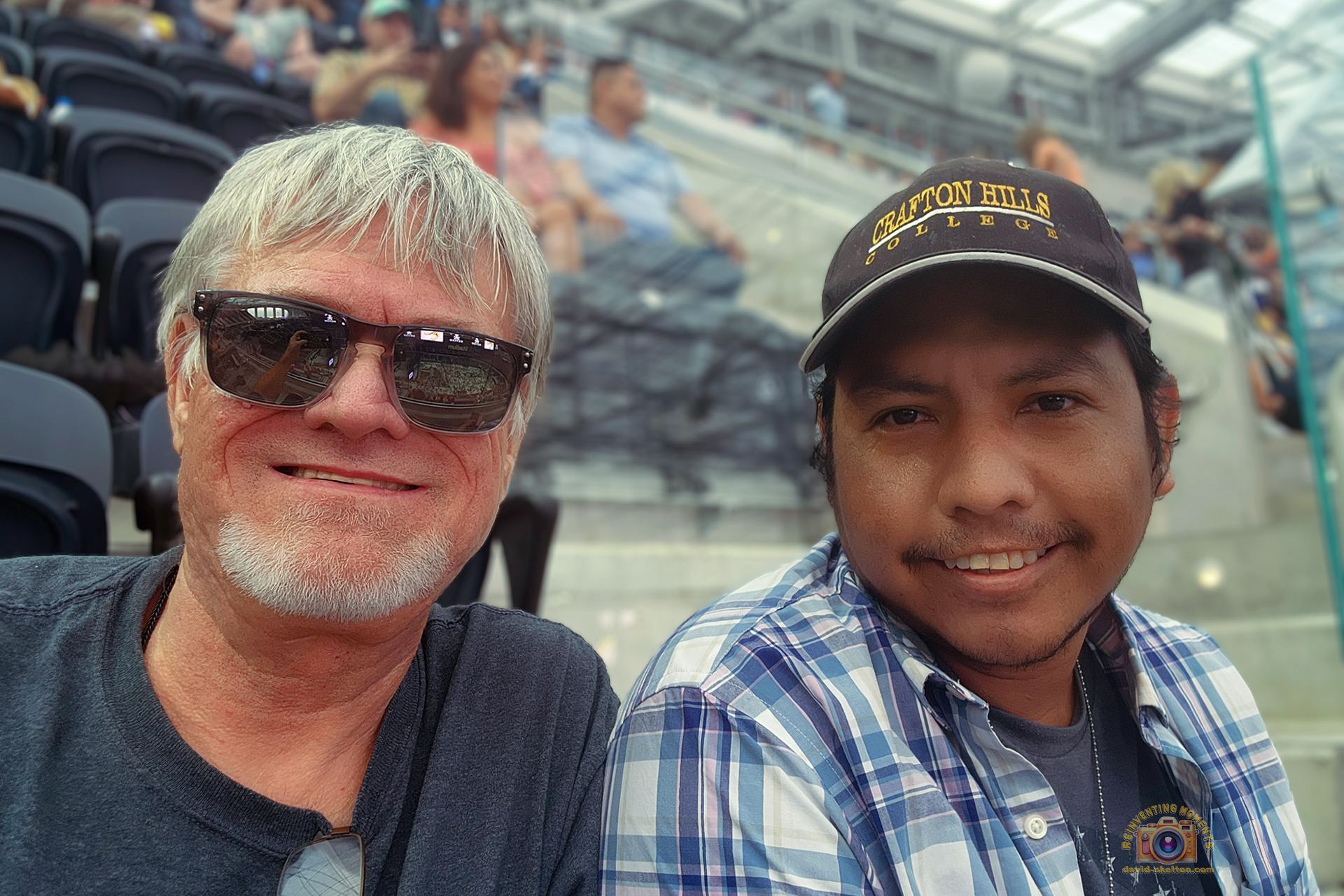 A man with white hair and sunglasses smiles next to his husband, who is wearing a plaid shirt and a baseball cap, sitting in the stands of a large, modern stadium.