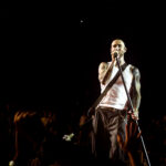 Frontman Adam Levine, in a white tank top showing his tattooed arms, sings passionately into a microphone on a dimly lit stage at the Acrisure Arena, with the hands of the audience visible in the foreground.