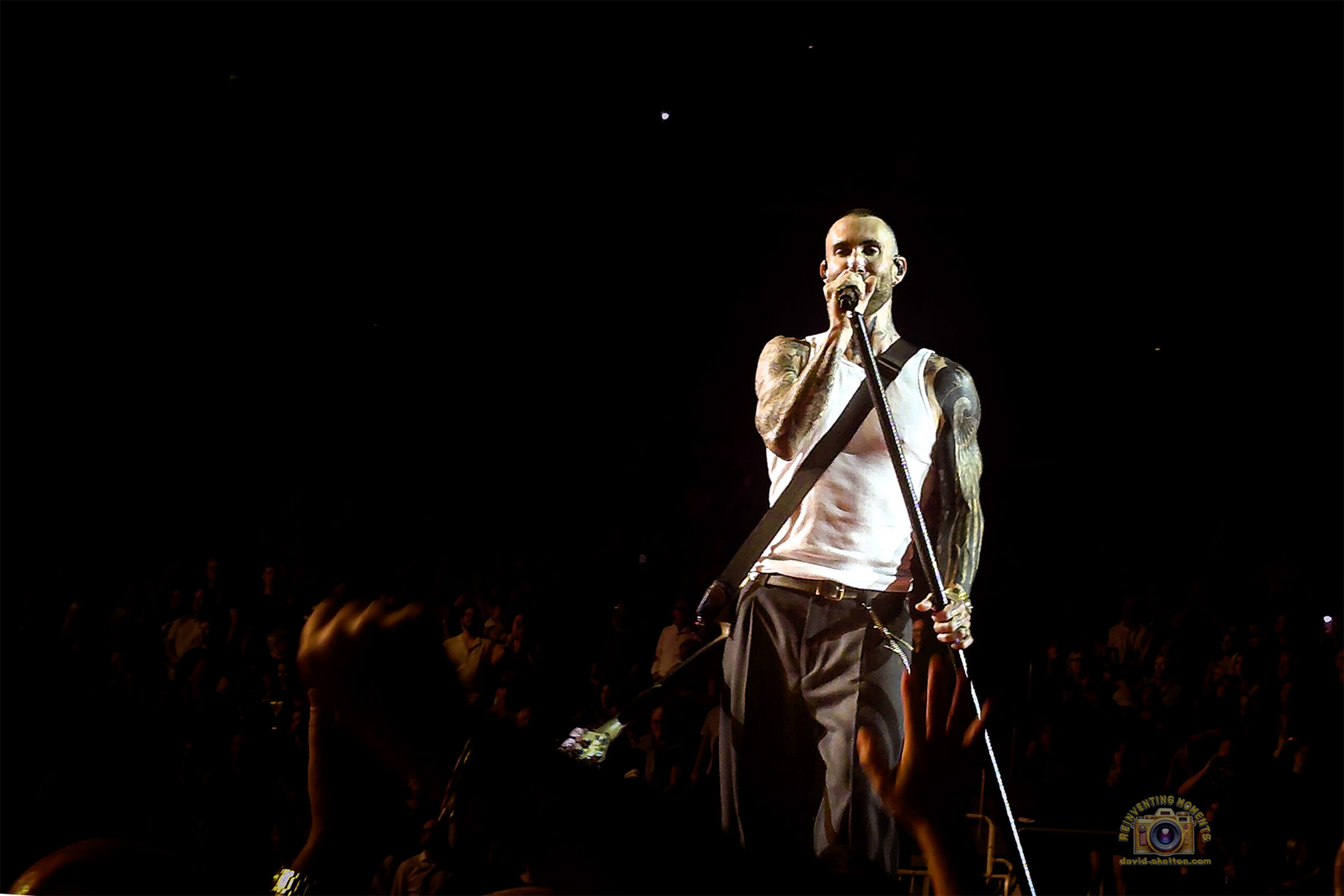 Frontman Adam Levine, in a white tank top showing his tattooed arms, sings passionately into a microphone on a dimly lit stage at the Acrisure Arena, with the hands of the audience visible in the foreground.
