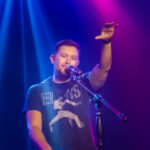 Country singer Scotty McCreery sings into a microphone on stage, his right hand raised, illuminated by dramatic blue and magenta spotlights.