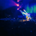 A wide stage view of Brad Paisley performing on the Weekend Warrior Tour, with a large video screen showing him playing guitar and the stage lit in blue