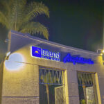 Close-up, low-angle night photo of the white textured building entrance sign for the North Island Credit Union Amphitheatre, with bright blue illuminated lettering and backlit palm trees overhead.