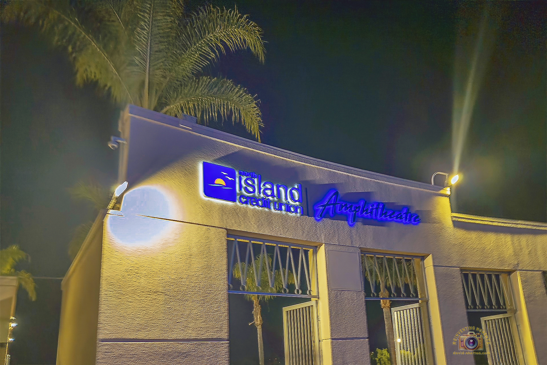 Close-up, low-angle night photo of the white textured building entrance sign for the North Island Credit Union Amphitheatre, with bright blue illuminated lettering and backlit palm trees overhead.