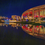 Vibrant, dark, wide-angle photo of the YouTube Theater and curving SoFi Stadium exterior at night, with the structures’ colorful lights reflecting sharply on the smooth, dark water of the lake in the foreground.