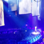 Wide shot of Carrie Underwood on stage at STAPLES Center under dramatic blue and purple lights, raising her arm during a performance of "Jesus Take The Wheel." Her face is visible on a large video screen to the left.