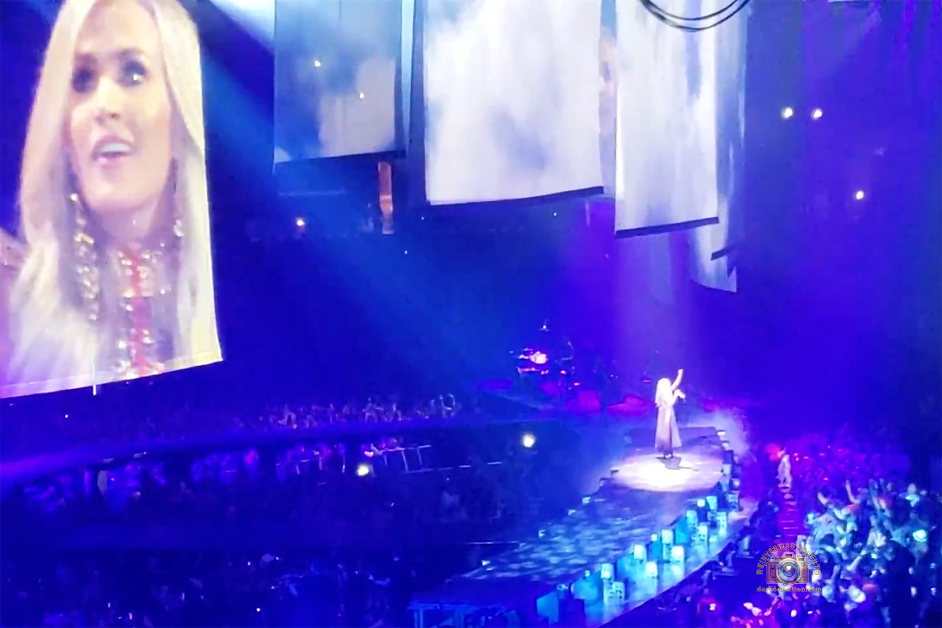 Wide shot of Carrie Underwood on stage at STAPLES Center under dramatic blue and purple lights, raising her arm during a performance of "Jesus Take The Wheel." Her face is visible on a large video screen to the left.