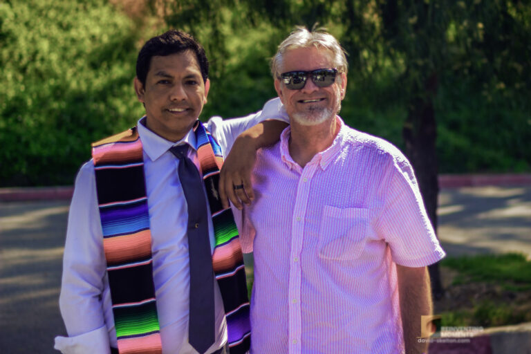 A sunny outdoor portrait of Gio Skelton wearing a colorful striped serape graduation sash and tie, resting his arm affectionately on the shoulder of his husband, David Skelton, who is wearing sunglasses and a pink short-sleeve button-down shirt.
