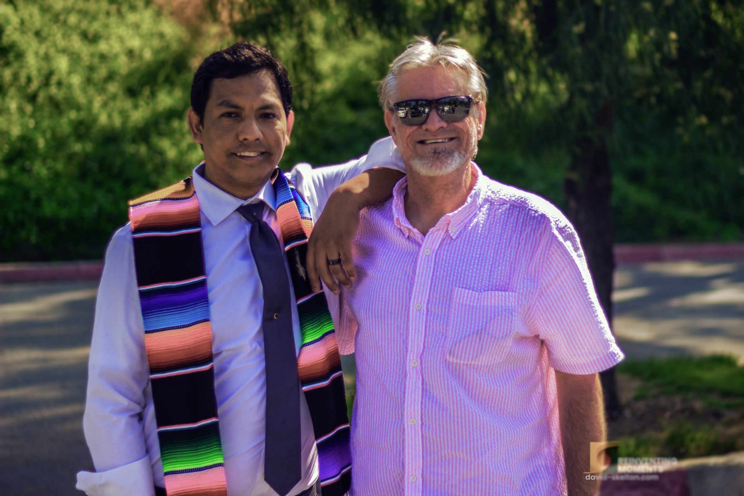 A sunny outdoor portrait of Gio Skelton wearing a colorful striped serape graduation sash and tie, resting his arm affectionately on the shoulder of his husband, David Skelton, who is wearing sunglasses and a pink short-sleeve button-down shirt.