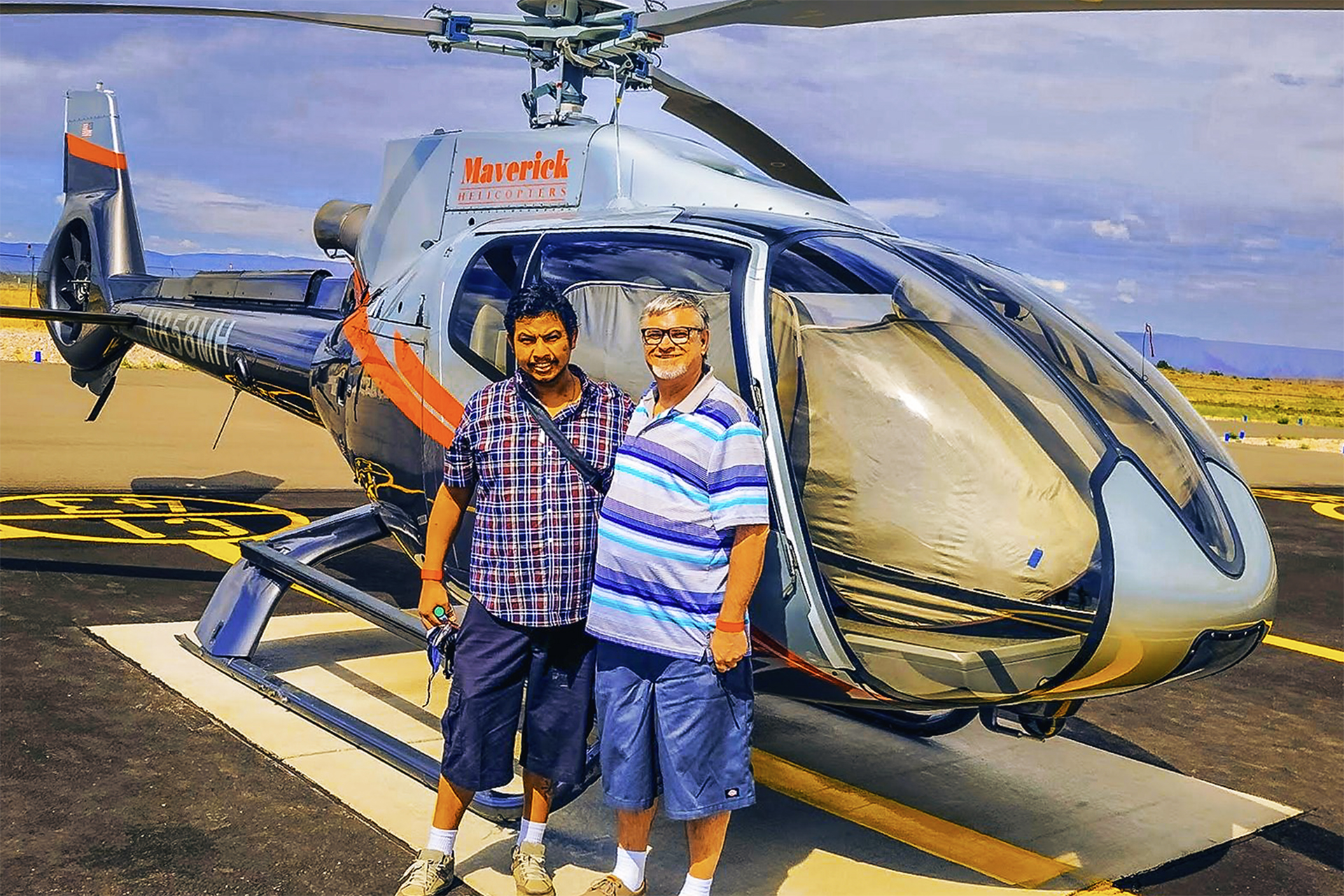 David and Gio standing on the tarmac at Grand Canyon West in front of a silver Maverick Helicopters Airbus EC130 ECO-Star, smiling during their Las Vegas to Grand Canyon tour.