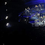 A concert scene featuring a large video screen on the left showing a close-up of Eric Church singing into a microphone while wearing sunglasses and an American flag scarf. On the right, the full stage setup is visible with backing vocalists and the band performing under blue lights, with the artist standing stage-right.