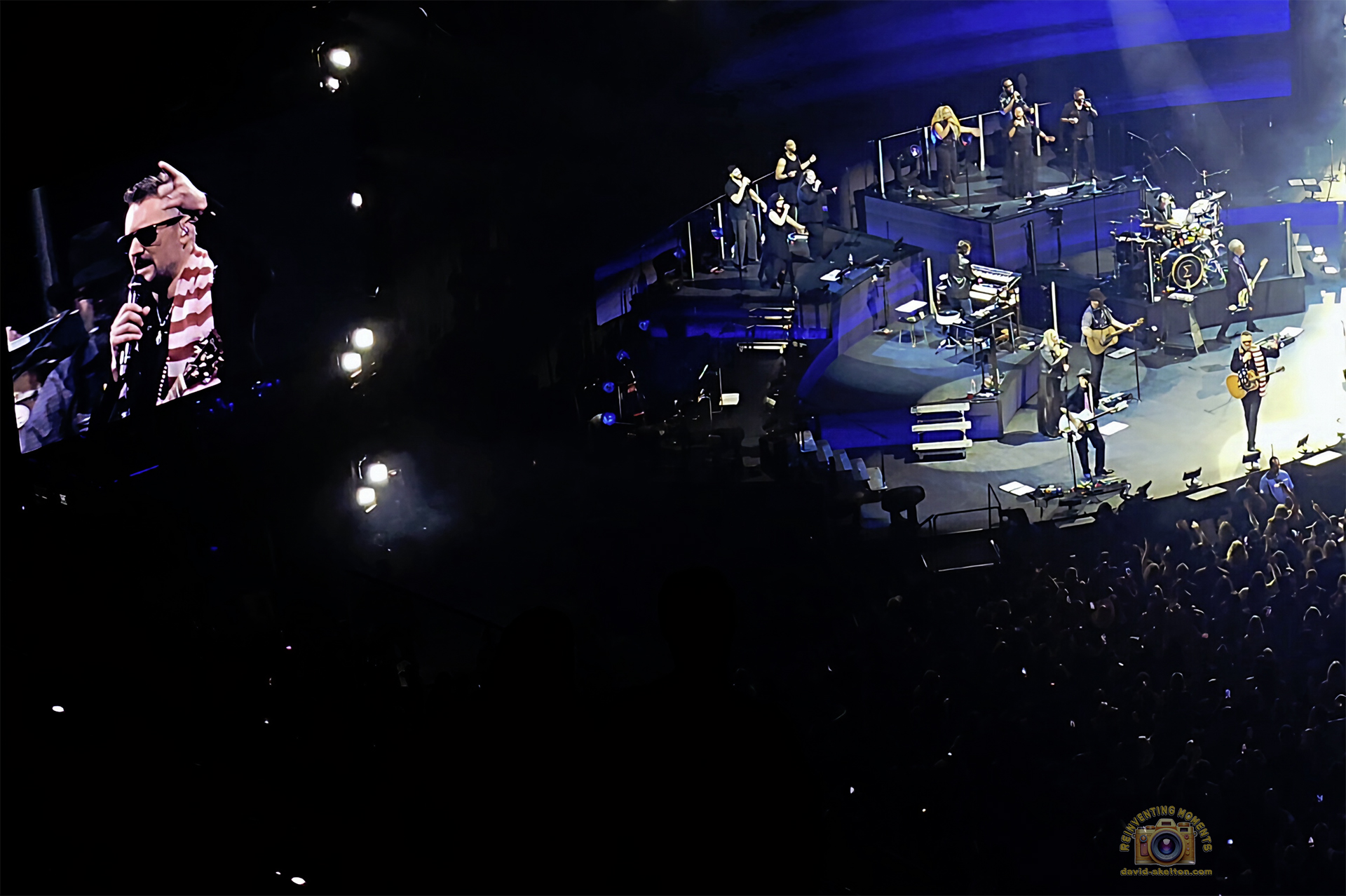 A concert scene featuring a large video screen on the left showing a close-up of Eric Church singing into a microphone while wearing sunglasses and an American flag scarf. On the right, the full stage setup is visible with backing vocalists and the band performing under blue lights, with the artist standing stage-right.