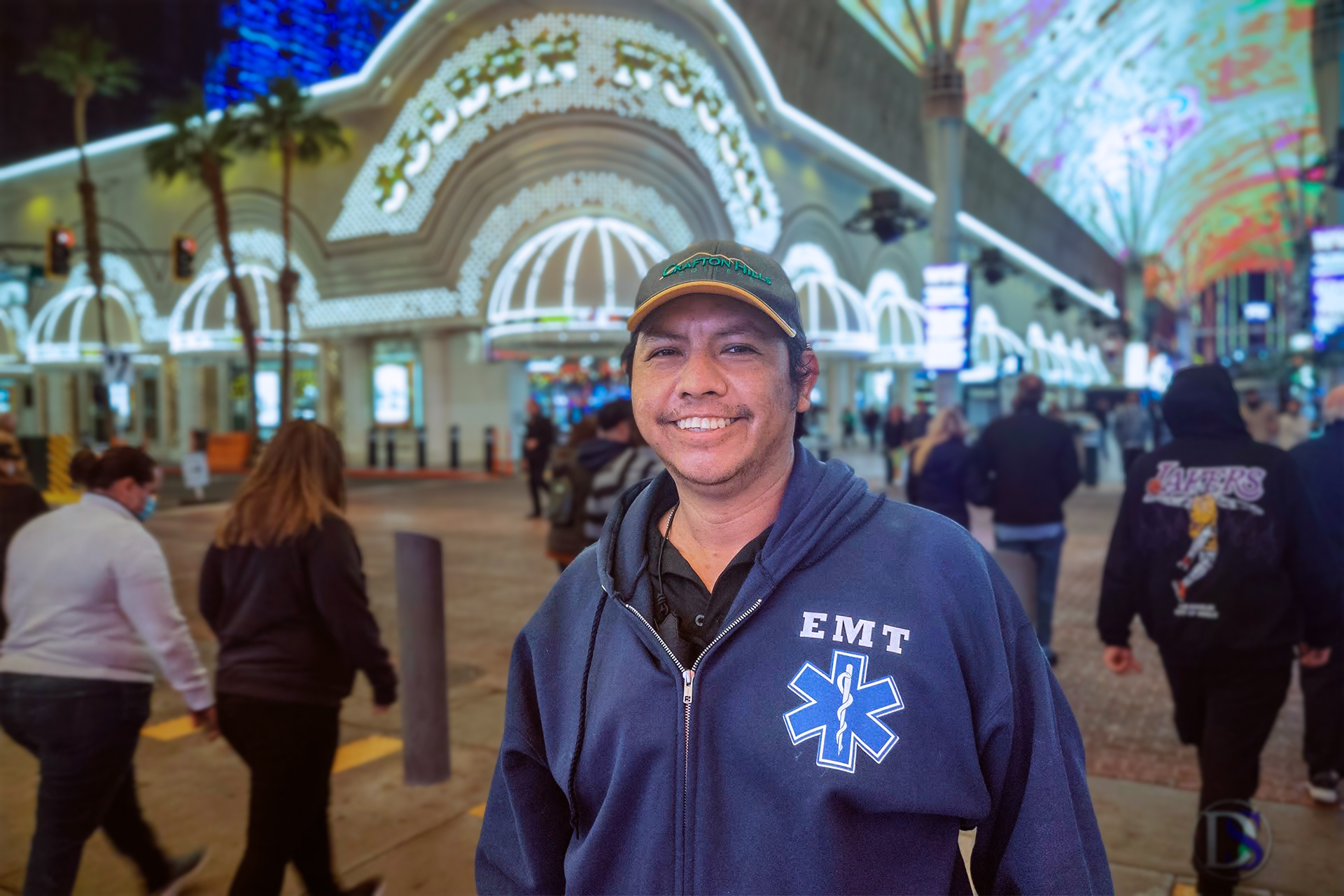 Gio Skelton smiling comfortably wearing a blue EMT hoodie and baseball cap, standing in the center of a pedestrian walkway in front of the brightly illuminated Golden Nugget casino entrance on Fremont Street at night.